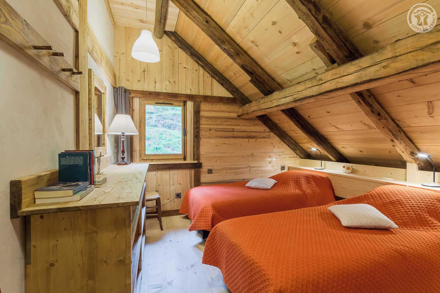 Une cabane chaleureuse avec deux lits, un bureau avec des livres, une fenêtre et un plafond en bois.
