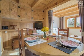A wooden interior with a dining table set for two, a television, and a window with curtains.