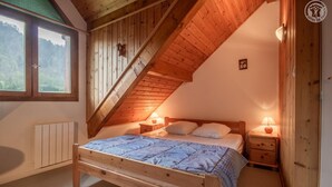 A bedroom with a sloped ceiling, wooden walls, a bed with a blue quilt, two bedside tables with lamps, and a window with a view of trees. - LA CASCADE - SAINT PANCRACE (SAINT PANCRACE)