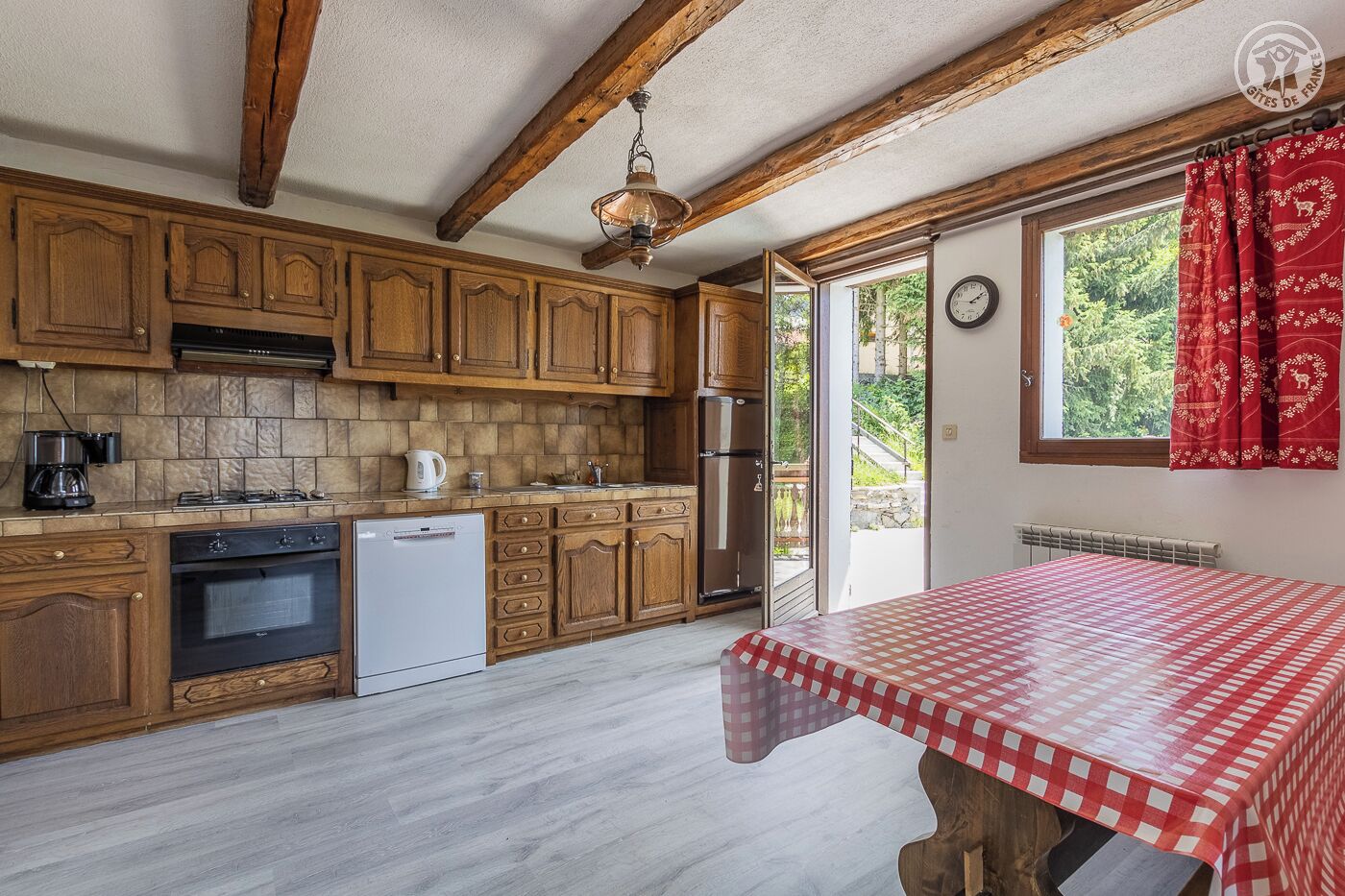 A kitchen with wooden cabinets, a dishwasher, a stove, and a refrigerator. There is a window with red curtains and a view of greenery outside.