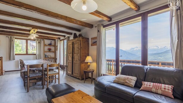 Una sala de estar acogedora con un sofá de cuero, una mesa de centro de madera y un comedor con una mesa y sillas de madera. Amplias ventanas ofrecen vistas a la montaña.