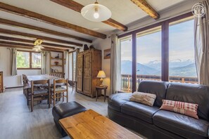 Ein gemütliches Wohnzimmer mit einer Ledersofa, einem Holztisch und einem Essbereich mit Tisch und Stühlen. Große Fenster bieten Blick auf die Berge.