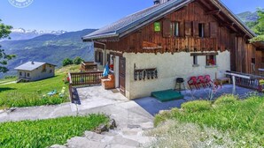 Un chalé rústico con una escalera de piedra que lleva a una puerta de madera, un pequeño porche con un banco y una vista de montañas nevadas al fondo.
