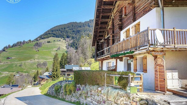 En traditionel schweizisk chalet med en træbalkon, stenvæg og udsigt over en grøn dal og skovklædte bakker.
