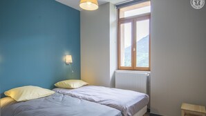 A bedroom with two beds, a window with a view of mountains, a radiator, and a small wooden stool.