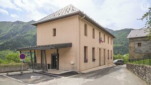 A beige building with a covered entrance, a parking area, and a mountain backdrop.