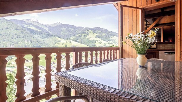 Ein Holzbalkon mit Glastisch und Korbstühlen mit Blick auf eine gebirgige Landschaft.