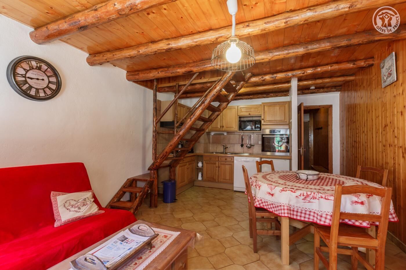 Un salon chaleureux avec un canapé rouge, une table à manger en bois et un escalier menant à la cuisine.