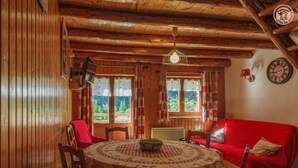Una habitación acogedora con paredes y techo de madera, una mesa redonda con mantel floral, un sofá rojo y dos ventanas con cortinas.