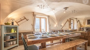 Un comedor rústico con una mesa de madera preparada para una comida, un banco y una estantería con vajilla.