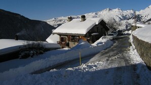 A snow-covered mountain road with a rustic building, a parked car, and a clear blue sky.