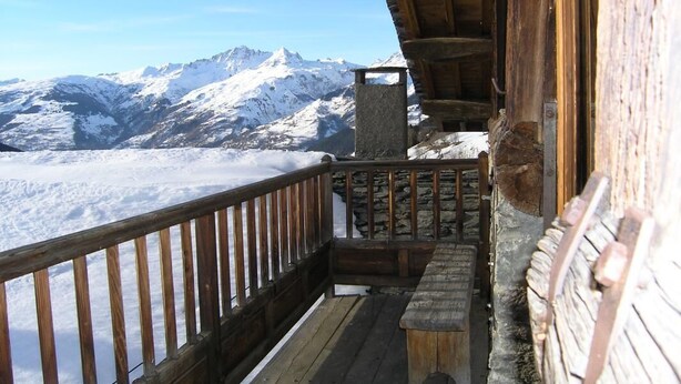A wooden balcony with a stone bench and a stone fireplace, overlooking a snowy mountain range.