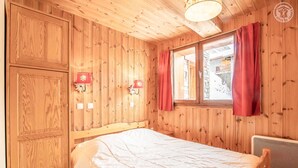 Une pièce en bois avec un lit, deux lampes fixées au mur et une fenêtre ornée de rideaux rouges.