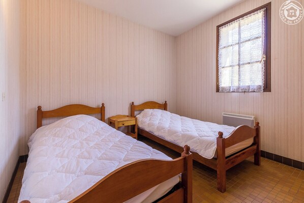 Una habitación con dos camas de madera, una mesita de noche pequeña, un radiador y una ventana con cortinas de encaje.