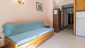 A room with a wooden sofa, a small table, a kitchen area with a refrigerator and stove, and a door leading to another room. - LE PRESBYTERE 2 - MONTSAPEY (MONTSAPEY)