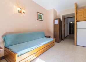 A room with a wooden sofa, a small table, a kitchen area with a refrigerator and stove, and a door leading to another room. - LE PRESBYTERE 2 - MONTSAPEY (MONTSAPEY)
