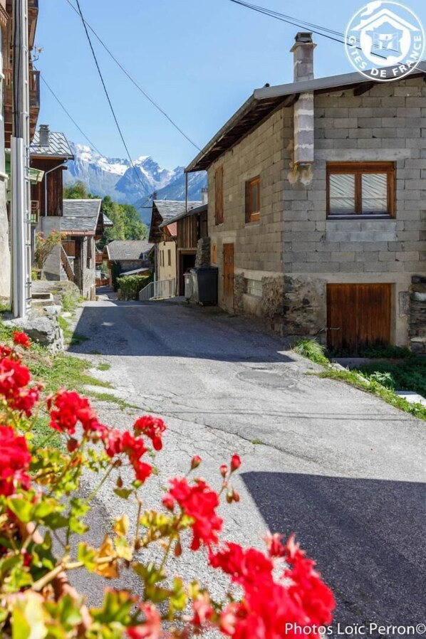 A narrow street in a mountainous village with stone buildings and a clear view of snow-capped mountains in the background.