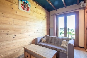A wooden cabin interior with a sofa, a small table, and a window with a view of greenery.