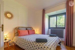 A bedroom with a patterned bedspread, a bedside lamp, a window with curtains, and a radiator.