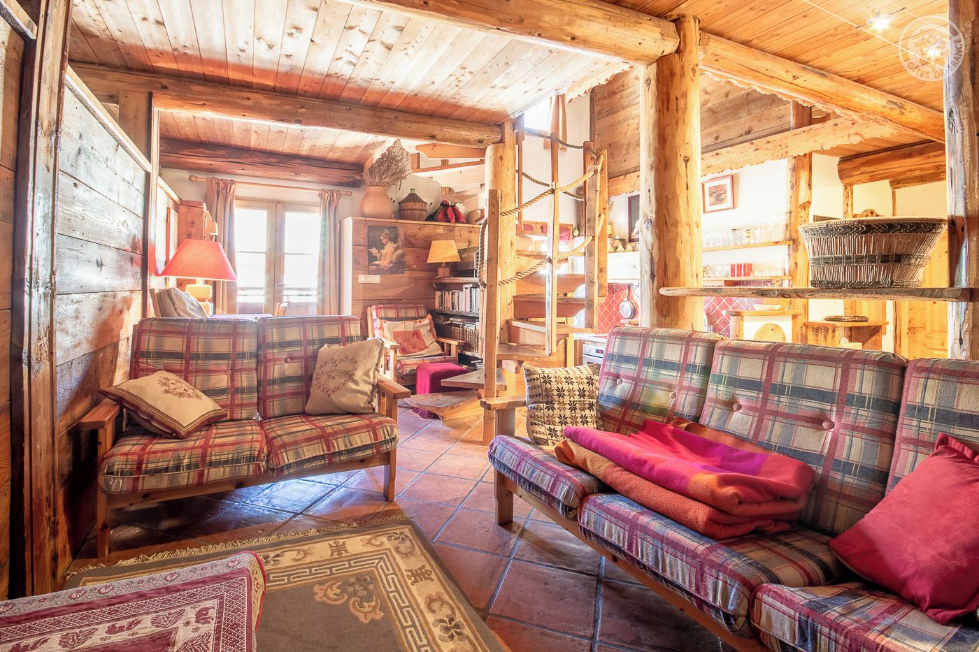 A cozy log cabin interior with plaid furniture, a fireplace, and a rustic wooden ceiling.