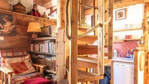 A rustic wooden interior with a loft bed, shelves with books, a kitchen area, and a hanging rope.