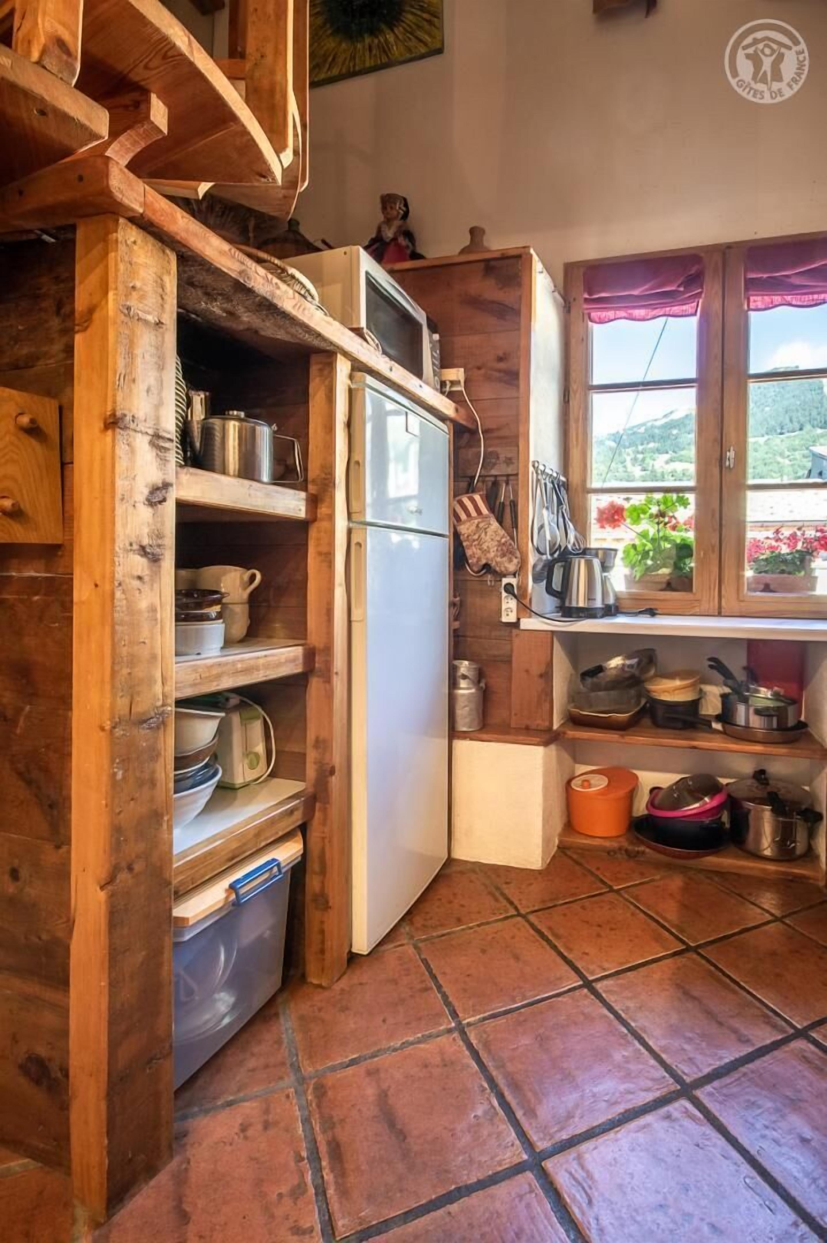 A rustic kitchen with wooden shelves, a refrigerator, and a window with flowers.