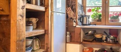 A rustic kitchen with wooden shelves, a refrigerator, and a window with flowers.