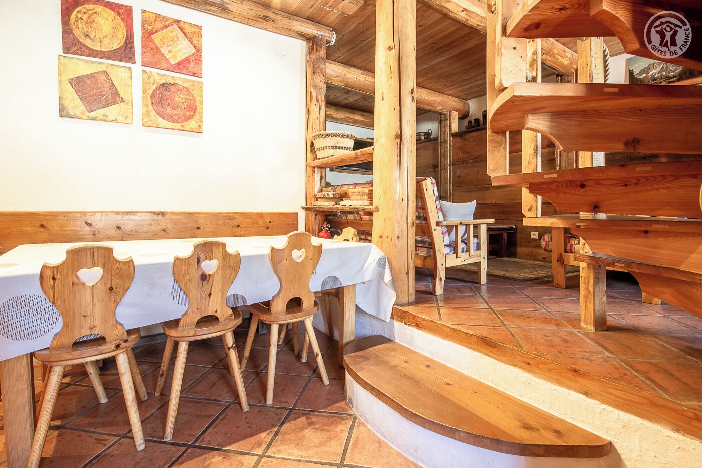 A rustic wooden interior with a long table and chairs, a staircase leading to an upper level, and a wall with framed artwork.