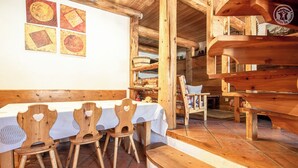 A rustic wooden interior with a long table and chairs, a staircase leading to an upper level, and a wall with framed artwork.