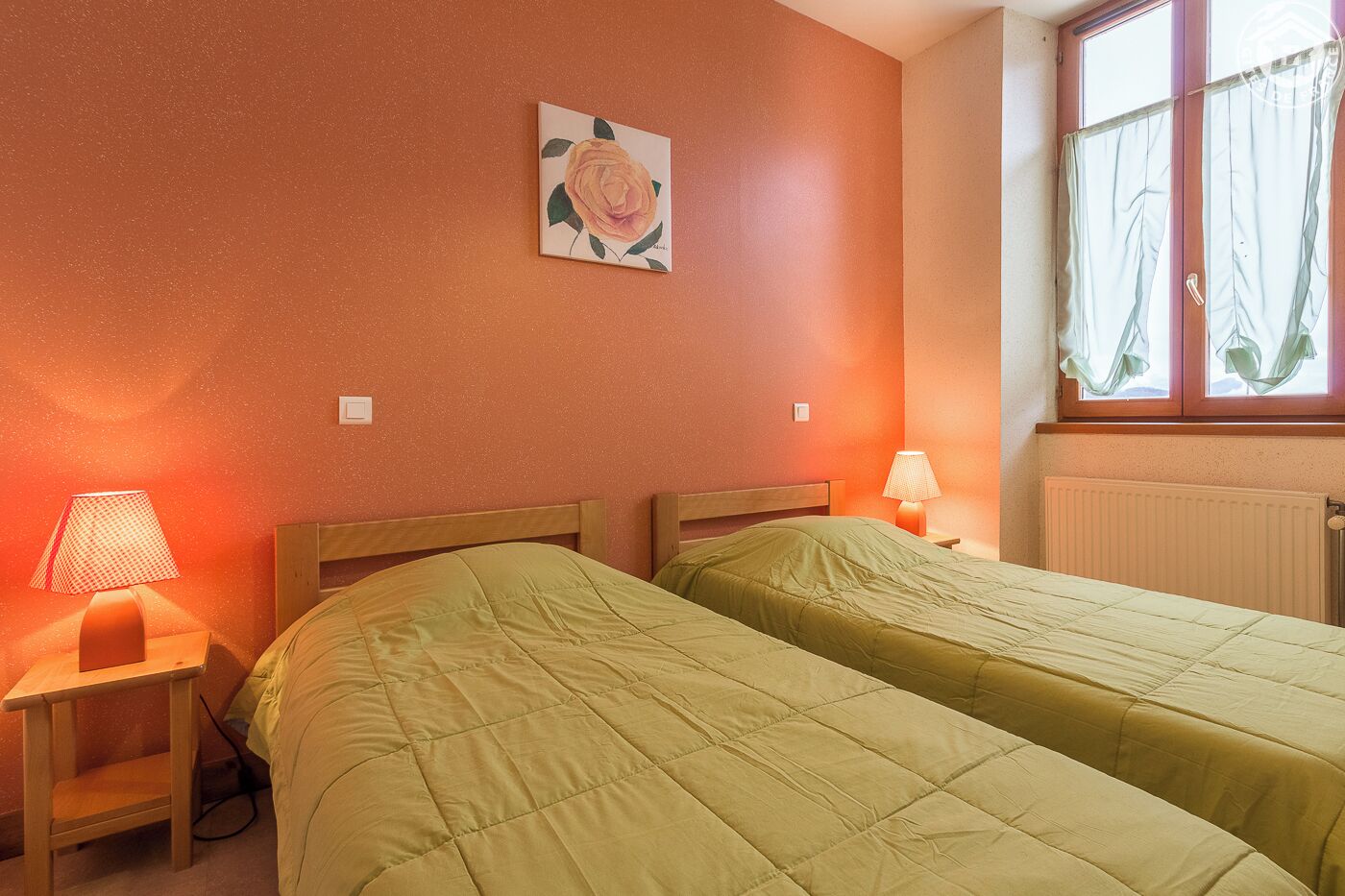 A room with two beds, each with a green bedspread, a nightstand with a lamp, and a painting of a rose on the wall.