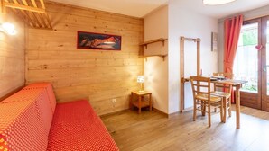 A cozy room with a wooden wall, a bed with a red patterned bedspread, a small table with a lamp, and a dining area with chairs and a table. - LACHAT - CHALET LA LICORNE (NOTRE-DAME-DE-BELLECOMBE)