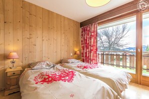 A bedroom with two beds, wooden walls, a balcony with a view of trees and mountains, and a window with red curtains.