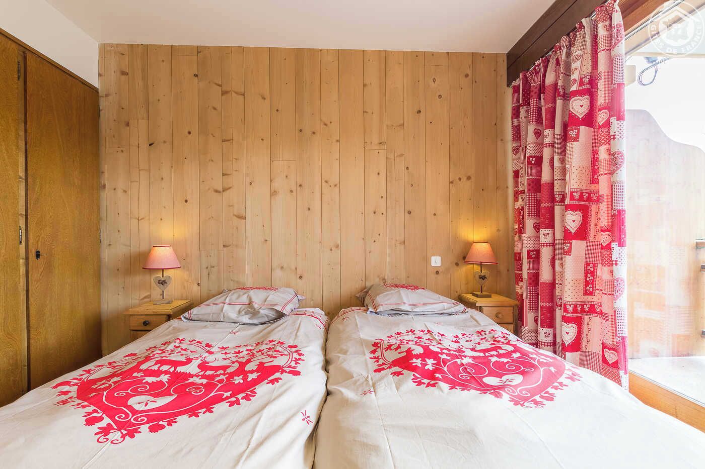 A room with two beds, wooden panel walls, and a window with patterned curtains.