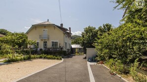 Une maison à deux étages avec un balcon, une allée et un jardin agrémenté de diverses plantes et d’un petit abri de jardin.