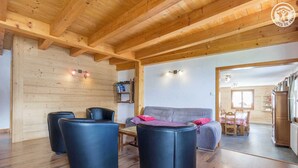 Un salon chaleureux avec un plafond en bois, un canapé gris, des fauteuils noirs et un coin repas comprenant une table et des chaises.