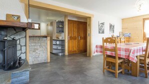 Une pièce chaleureuse avec une cheminée en pierre, des étagères en bois, une table à manger recouverte d’une nappe à carreaux et une porte en bois.