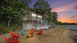 A beachfront property with a deck, red and blue Adirondack chairs, a fire pit, and a view of the ocean. - 🌅 Private Beach | Sunset Bay Views | House+Camper | M22 ➠ 7633 (Traverse City)