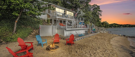 A beachfront property with a deck, red and blue Adirondack chairs, a fire pit, and a view of the ocean.