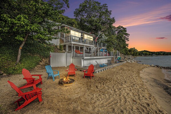 A beachfront property with a deck, red and blue Adirondack chairs, a fire pit, and a view of the ocean.