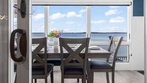 A dining area with a table set for four, a vase with flowers, and a view of the sea through large windows. - 🌅 Private Beach | Sunset Bay Views | House+Camper | M22 ➠ 7633 (Traverse City)