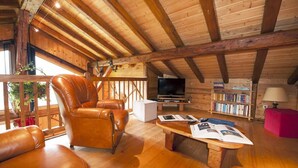 A cozy cabin interior with a wooden ceiling, leather recliner, a flat-screen TV, a bookshelf, and a dining table with magazines.