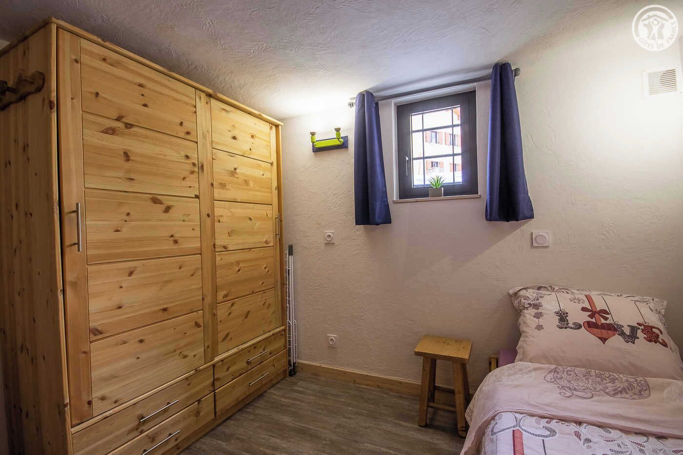 A bedroom with a wooden wardrobe, a small bedside table, a bed with patterned bedding, a window with curtains, and a small plant on the windowsill.