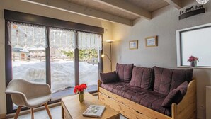 Une pièce chaleureuse avec un canapé en bois, un fauteuil, une table sur laquelle repose un livre et une fenêtre ornée de rideaux en dentelle.