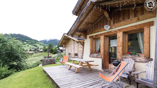 Eine Holzhütte mit einer Terrasse, Sitzgelegenheiten im Freien und Blick auf ein Tal.