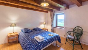 Ein Schlafzimmer mit Holzbalkendecke, einem Bett mit blauen, gemusterten Bettdecken, zwei Nachttischlampen, einem Fenster mit Vorhängen und einem grünen Sessel.