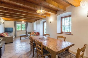 Un vaste espace de vie comprenant une table à manger en bois, des sièges confortables, un téléviseur et de grandes fenêtres laissant entrer la lumière naturelle.