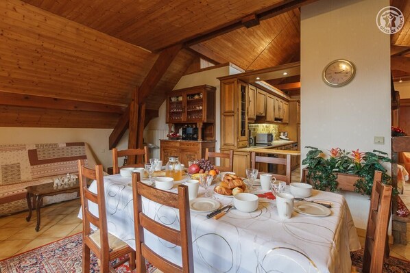 A dining room with a wooden table set for breakfast, wooden chairs, and a kitchen in the background.