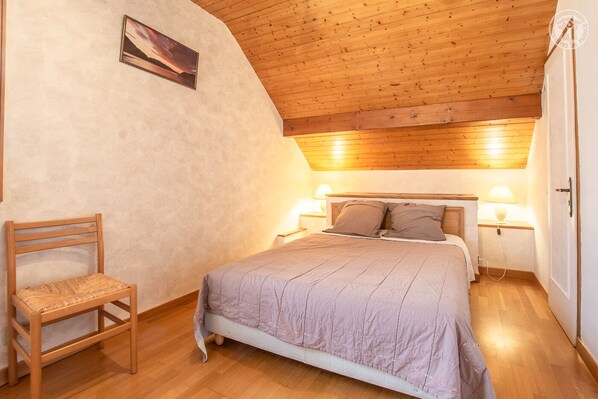 A bedroom with a wooden ceiling, a bed with a headboard, two bedside lamps, a chair, and a painting on the wall.