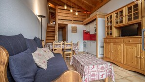 A cozy living room with a sofa, a dining table, and a kitchen area with wooden cabinets and a TV. - CRISTAL 4 - HAUTELUCE LES SAISIES (HAUTELUCE LES SAISIES)