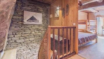 Une pièce rustique avec un mur en pierre, des poutres en bois, un lit superposé et un escalier doté d’une rampe en bois.
